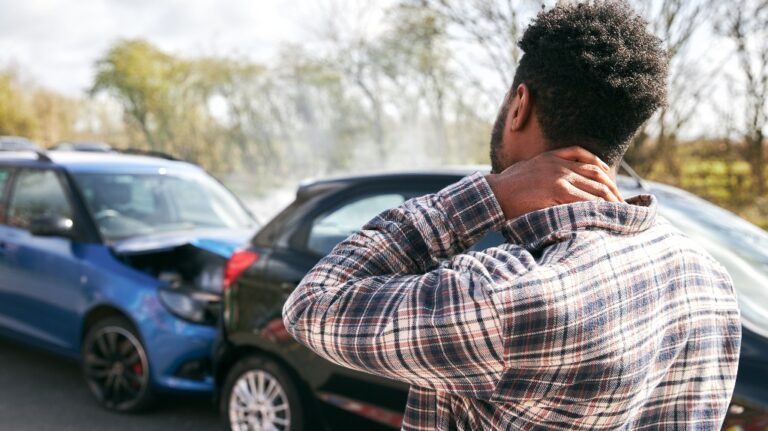 Young man rubbing neck in pain from whiplash injury standing by damaged cars