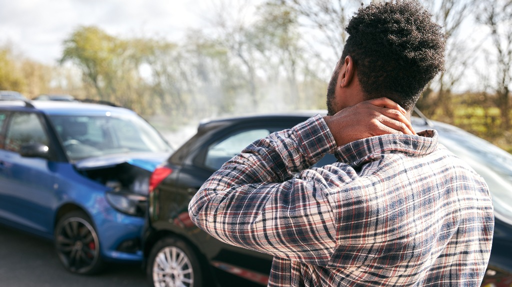 Young man rubbing neck in pain from whiplash injury standing by damaged cars