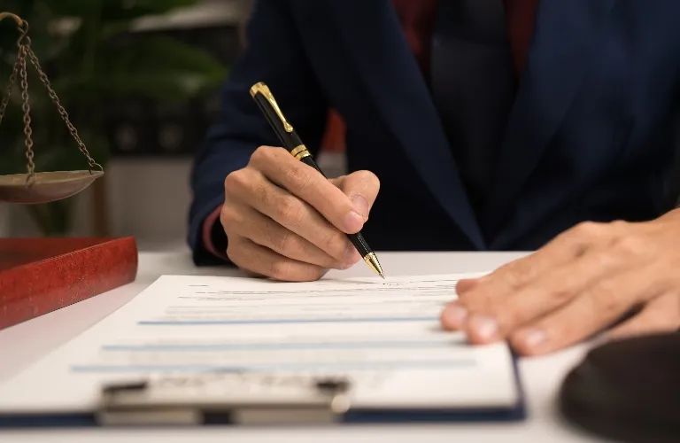 Person in dark suit signing legal documents with gold-accented pen at desk.