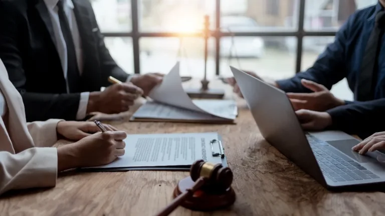 Lawyers reviewing documents and using a laptop at a wooden table.