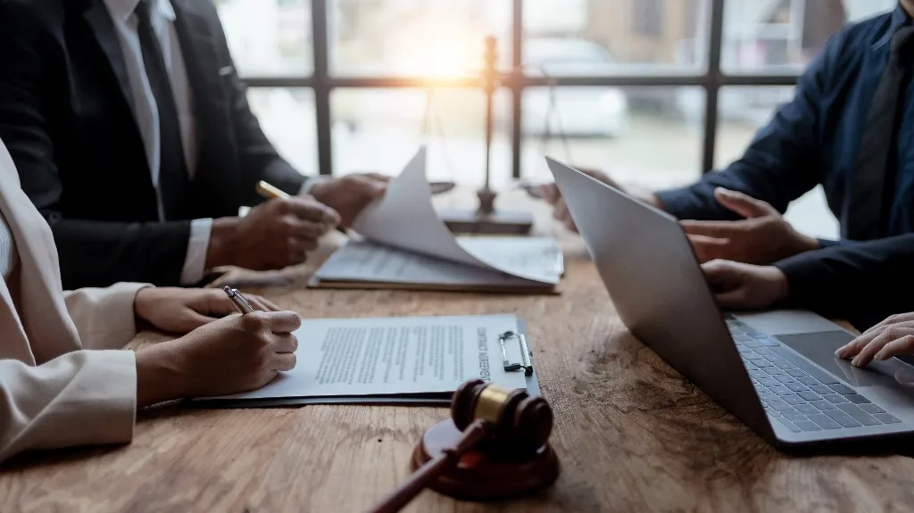 Lawyers reviewing documents and using a laptop at a wooden table.