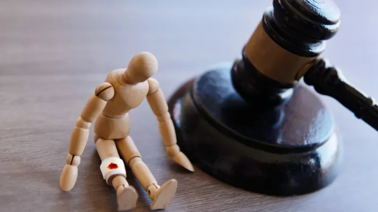 Wooden mannequin with a bloodied bandage on its leg next to a judge's gavel on a wooden surface.
