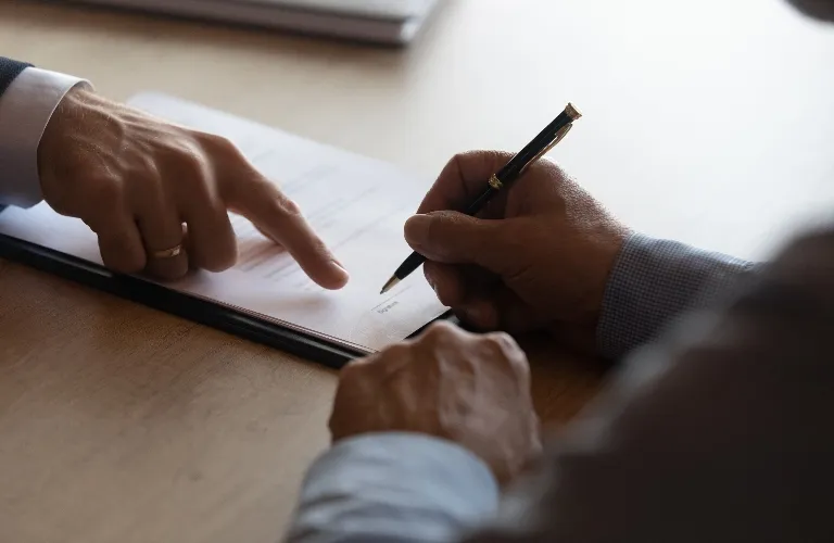 Two individuals engaged in signing a document on a wooden table, one pointing and the other holding a pen.