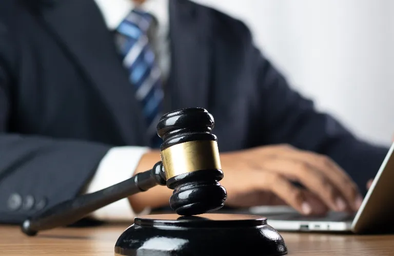Close-up of a black judge's gavel with a gold band on a wooden desk, blurred person in a suit working on a laptop in the background.