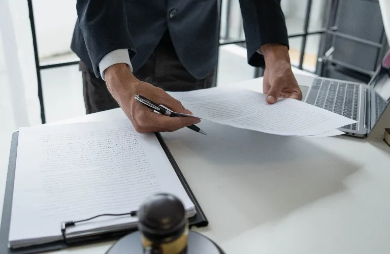 Person in dark suit reviewing and holding documents over a white desk with a laptop.