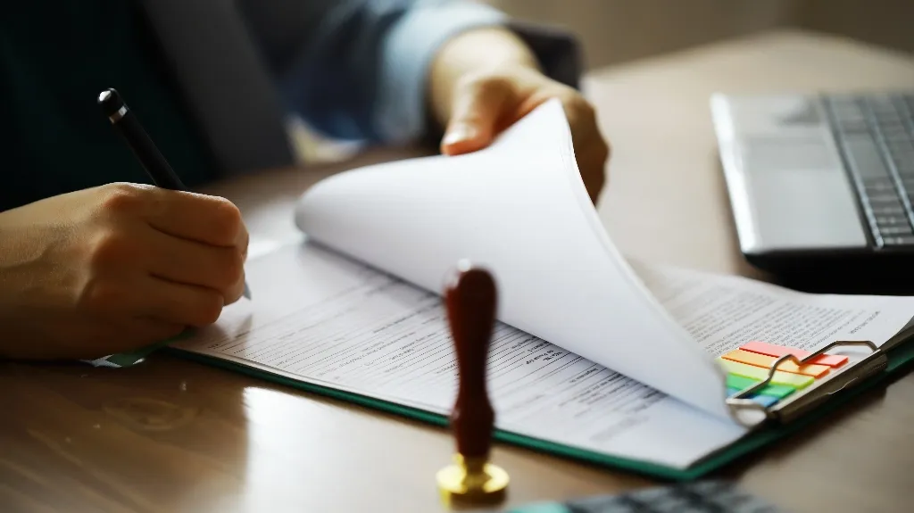 Person signing document on clipboard with colorful sticky tabs, laptop nearby on desk.