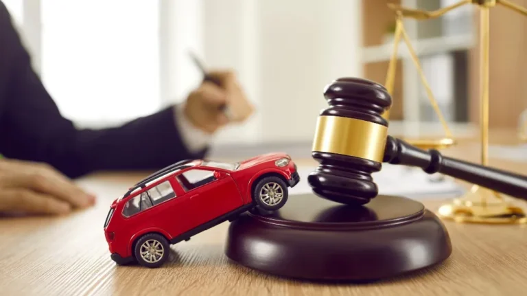 Red toy car leaning against wooden judge's gavel on desk with blurred person writing in background.