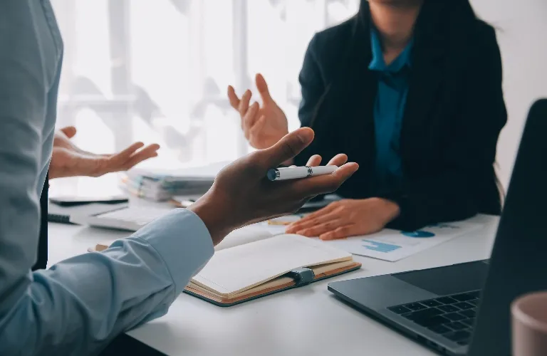 Two professionals engaged in a discussion with open notebook and laptop on white desk.