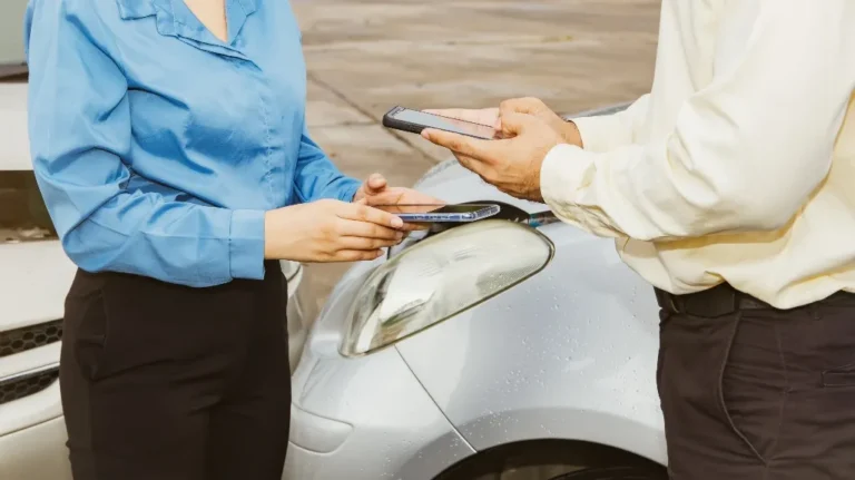 Two individuals exchanging smartphones near a silver car's headlight in a parking area.