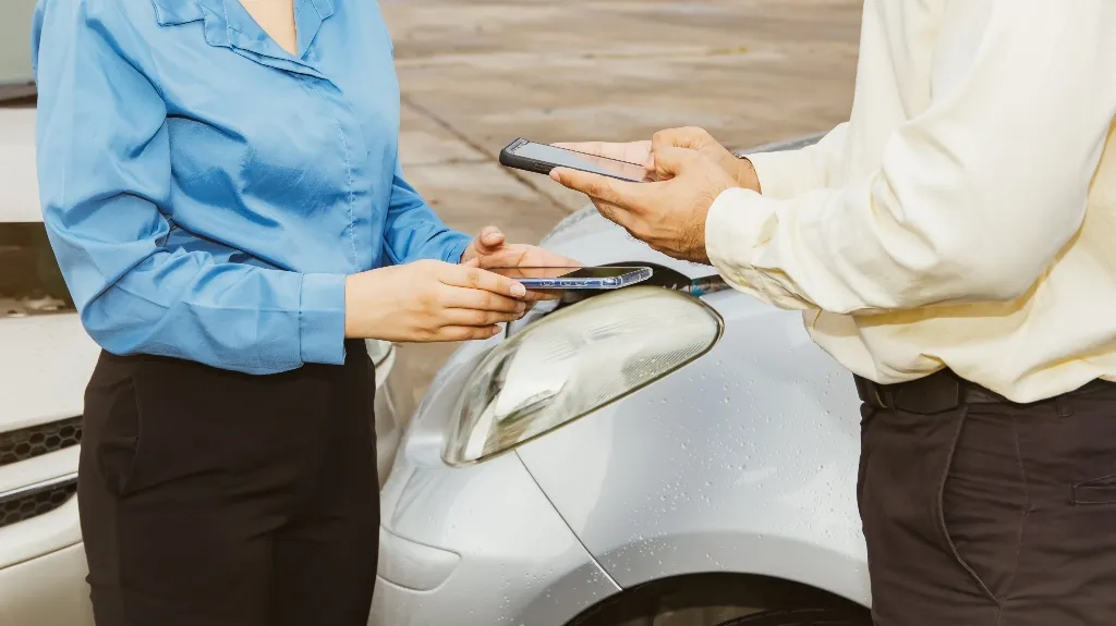 Two individuals exchanging smartphones near a silver car's headlight in a parking area.