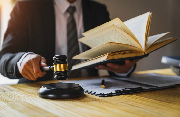 Person in suit holding open book and gavel over wooden desk with documents.