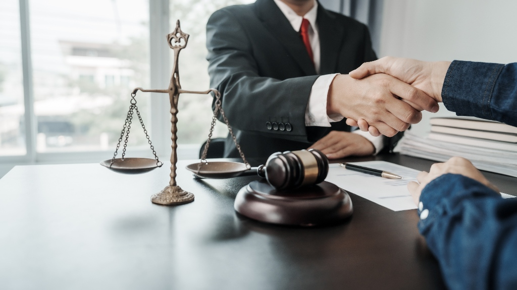 Two individuals shaking hands across a desk with a judge's gavel and scales of justice present.