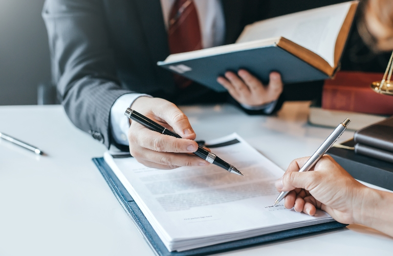 Two professionals reviewing and signing documents at a white desk, one holding a pen and the other a book.