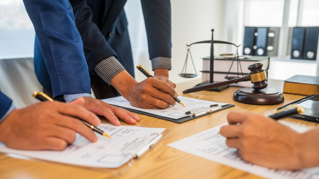 Three individuals in formal attire signing legal documents on clipboards, with a gavel and scales of justice on a wooden desk.