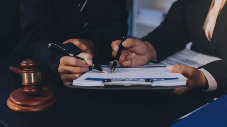 Two professionals in dark suits reviewing and signing a contract document, with a wooden gavel nearby.