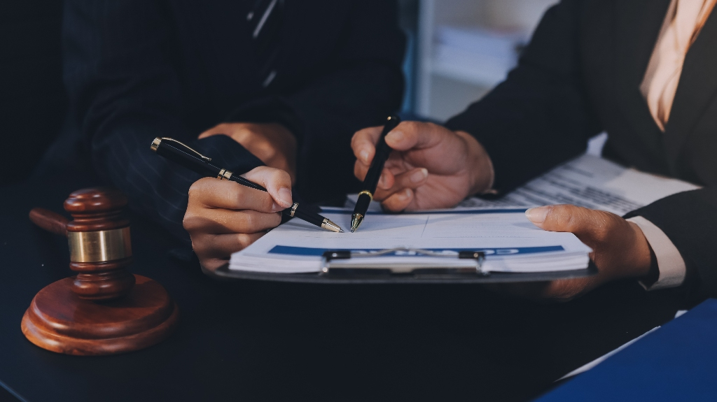 Two professionals in dark suits reviewing and signing a contract document, with a wooden gavel nearby.