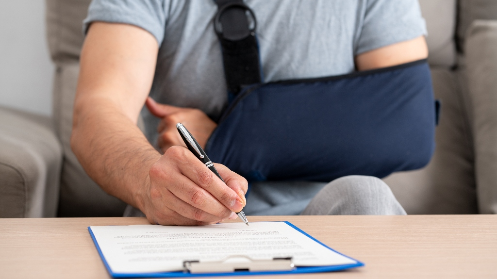 Person with arm in navy blue sling signing document on clipboard at table.