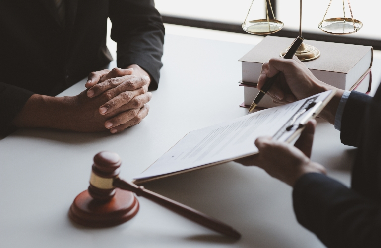 Two individuals in formal attire at a desk with legal documents, a gavel, and scales of justice.