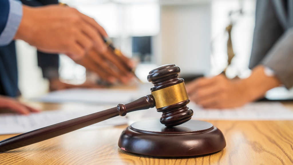 Wooden judge's gavel with brass band on desk, blurred hands signing documents in background.