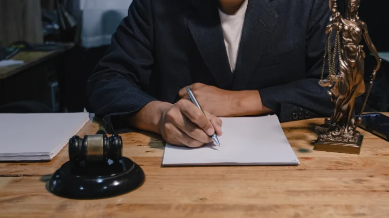 Person in dark suit writing on blank paper at wooden desk with gavel and Lady Justice statue.