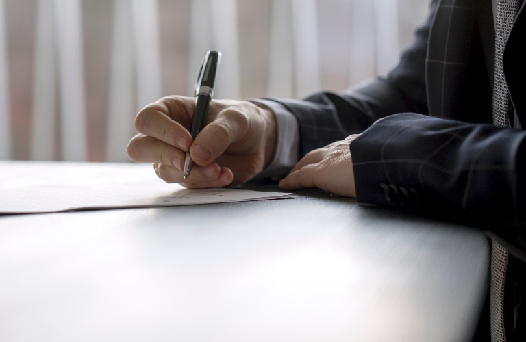 Close-up of a person in business attire writing with a pen on a document at a desk.