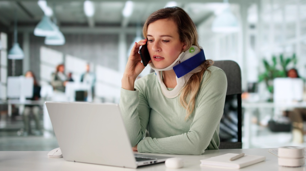 Woman wearing a neck brace talking on phone while working at a laptop in modern office.