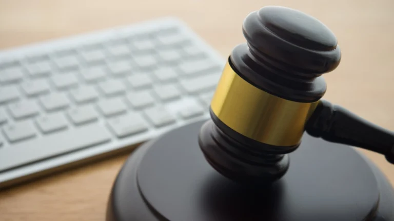 Close-up of a wooden judge's gavel with a gold band, placed near a white computer keyboard on a wooden desk.