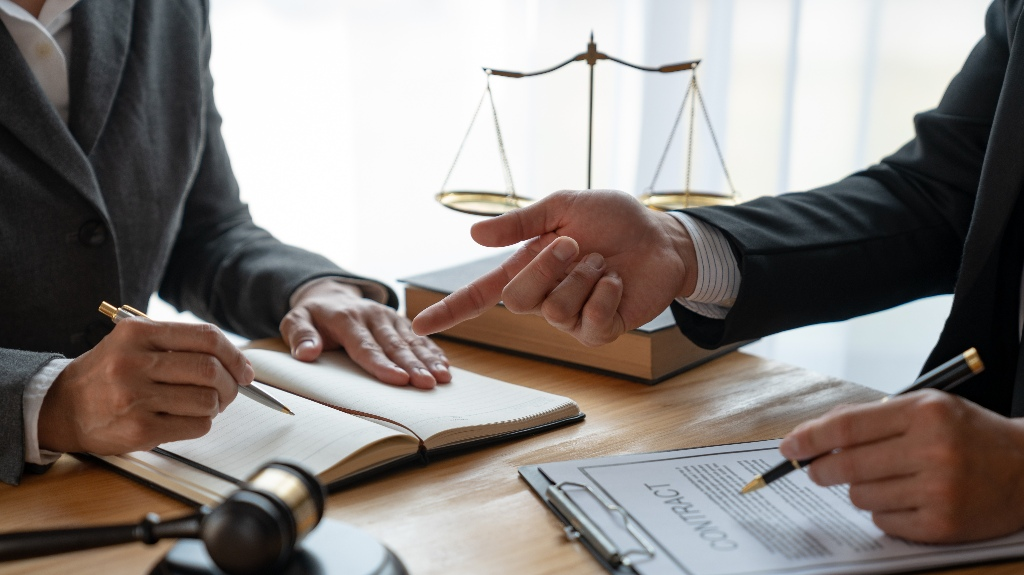 Two professionals in suits discussing a contract with legal scales and a gavel on the wooden desk.