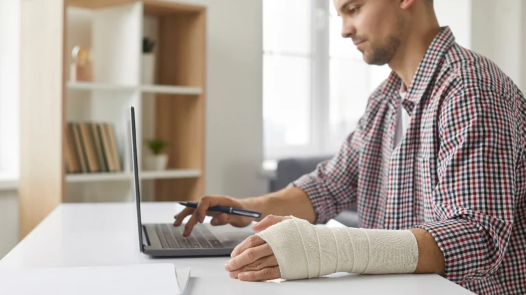Man with a bandaged left hand working on a laptop at a white desk in an office.