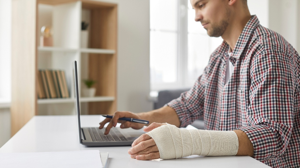Man with a bandaged left hand working on a laptop at a white desk in an office.
