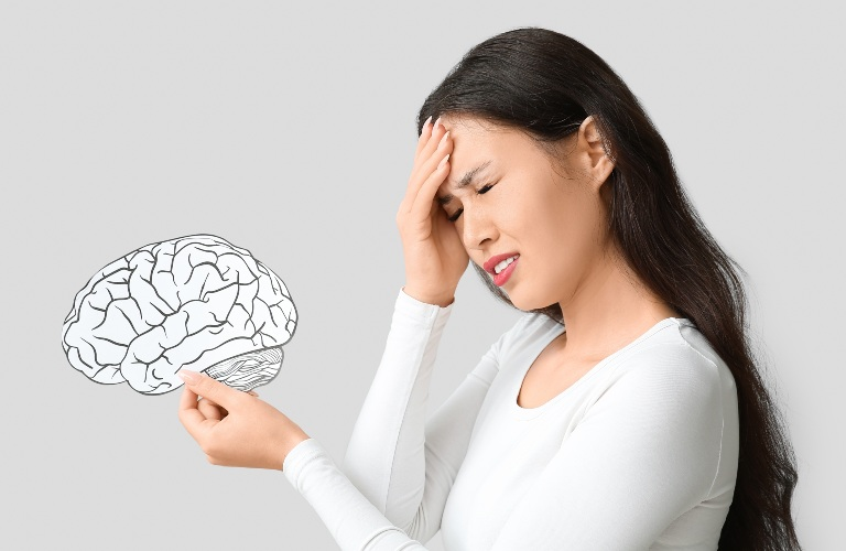 Woman in white shirt holding a brain illustration, touching forehead in discomfort.