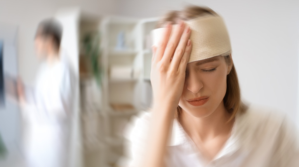Woman with a beige head bandage holding her head in pain, doctor blurred in background.