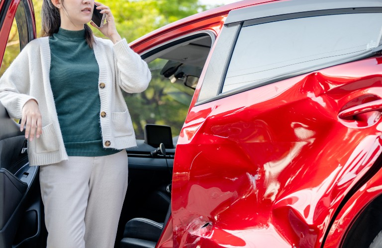 Woman in green top and beige cardigan making phone call beside red car with severe side door damage.
