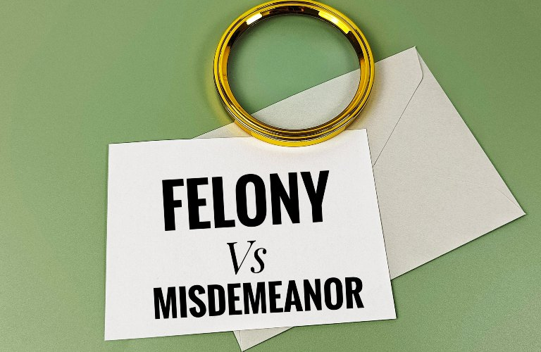 White card with bold black text "FELONY Vs MISDEMEANOR" beside a gold handcuff on a green surface.