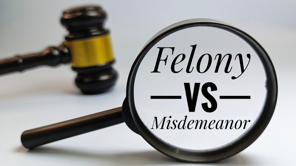 Magnifying glass focusing on text "Felony vs Misdemeanor" with a wooden judge's gavel in background.