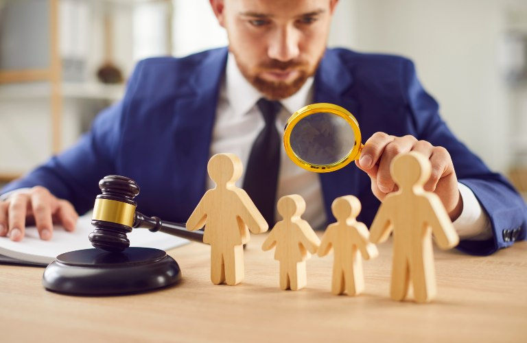 Man in blue suit examining wooden family figures with magnifying glass, gavel on table.