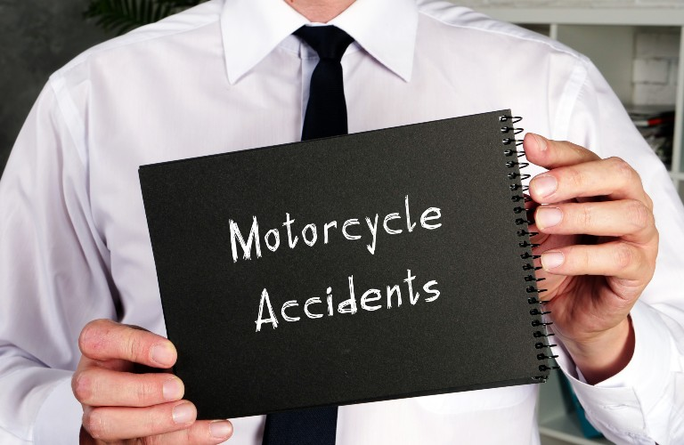 Man in white shirt and black tie holding black notebook with "Motorcycle Accidents" text.