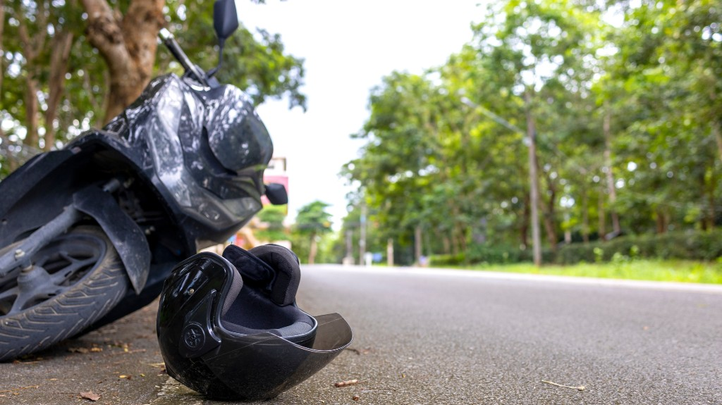 Black motorcycle helmet placed on the roadside near a parked black scooter with trees in the background.