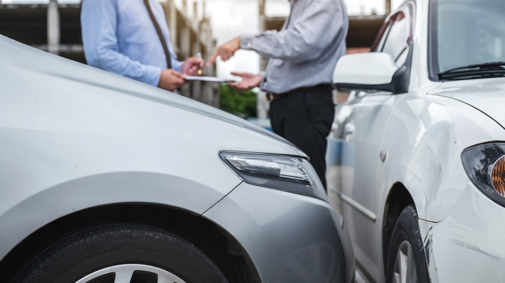 Two men in formal attire discussing beside two damaged white cars after a collision.